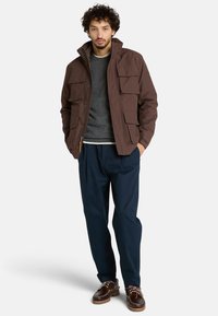 Giacca isolante marrone con quattro tasche frontali, colletto alto e chiusura con zip, abbinata a un maglione grigio e pantaloni blu scuro.
