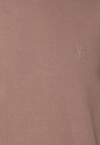 Camiseta rosa clara de tejido suave y texturizado con un pequeño diseño bordado de un cráneo con cuernos en el área del pecho izquierdo.