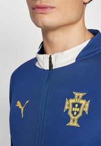 Giacca sportiva blu con colletto alto, realizzata in un tessuto testurizzato, con zip bianca e uno stemma giallo sul lato sinistro del petto raffigurante un logo di leone.