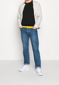 Sneakers bianchi, jeans blu, maglione nero sovrapposto a una camicia gialla e una giacca grigia chiara. L'outfit presenta un design casual e moderno.