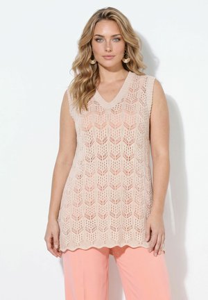Blond vrouw draagt een mouwloos beige gebreid topje met visgraatpatroon en perzikkleurige broek, staand tegen een witte achtergrond.