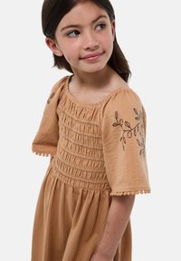Robe en coton beige avec de courtes manches à volants, corsage froncé et détails de feuilles brodés. L'ourlet présente un petit trim en pompons aux bords des manches.