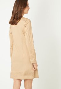 Mujer con cabello castaño hasta los hombros, vestida con un vestido beige de manga larga y hasta la rodilla, con detalles plisados en los puños y el dobladillo.