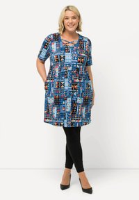 Lyhythihainen mekko, jossa on sininen monivärinen kangas, jossa on geometrisia muotoja, korostettuna rohkeilla väreillä ja sivutaskuilla, käytetään mustien legginssien päällä.