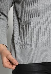Grå ribbad tröja med en strukturerad yta, med en framficka och en åtsittande midja. Tyget verkar mjukt och stretchigt.