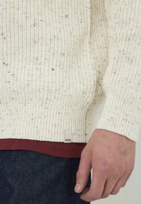 Pull en maille de couleur crème avec un motif texturé et des éclats de couleur. Manches à revers et ourlet côtelés, avec une petite étiquette BZB sur le côté.