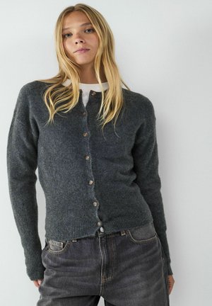 Grauer Wollcardigan mit Knopfleiste, geripptem Saum und Bündchen sowie Holzknöpfen. Getragen über einem weißen Hemd, kombiniert mit dunklen Jeans.