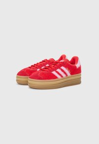 Par de sapatilhas Adidas Gazelle vermelhas com riscas brancas e solas platform cor de borracha, sobre um fundo cinza claro simples.
