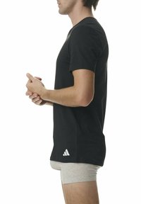 Svart bomullströja med korta ärmar, rund halsringning och en vit Adidas-logga på nedre vänstra sidan. Bärs med beiga shorts.