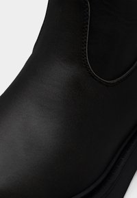 Κοντινή λήψη μύτης μαύρης δερμάτινης μπότες με ορατή ραφή και ελαστικό πλαϊνό πάνελ σε λευκό φόντο.