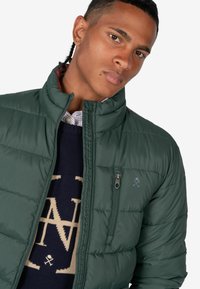 Chaqueta de plumas verde con cuello alto, textura acolchada y bolsillo en el pecho con cremallera. Lleva puesta sobre un suéter azul marino con un patrón ligero.