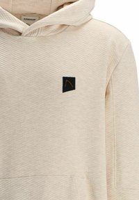 Sweat à capuche beige en tissu texturé. Présente une petite étiquette logo noire sur la poitrine, affichant un design minimaliste.