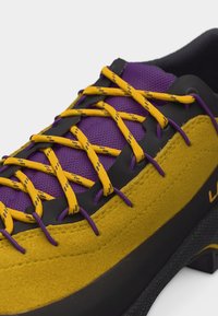 Κοντινό πλάνο ενός πεζοπορικού παπουτσιού σε κίτρινες και μωβ αποχρώσεις με κίτρινες κορδόνια, μαύρες τρύπες για τα κορδόνια και υφή στη σόλα, που αναδεικνύει το αναπνεύσιμο ύφασμα και το ανθεκτικό σχεδιασμό.