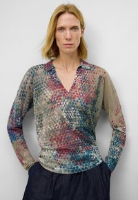 Chemisier à manches longues et col en V en tissu transparent, présentant un motif multicolore avec des points rouges, bleus et verts sur un fond beige.