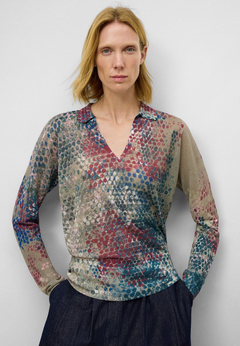 Chemisier à manches longues et col en V en tissu transparent, présentant un motif multicolore avec des points rouges, bleus et verts sur un fond beige.