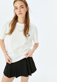 Witte katoenen T-shirt met een graphic van een hond met een koptelefoon, met de tekst "HOU VOL EN BLEEF GLUNDEREN!" in het blauw. Gecombineerd met een zwarte uitlopende rok.