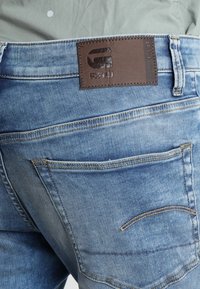 Ljusblå denimjeans med en blekt finish, som har en brun läderlapp med "G-STAR RAW"-logotyp och detaljerad söm vid fickorna.