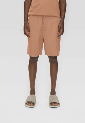 Mies, joka pukeutunut vaaleanruskeisiin naruilla säädettäviin shortseihin ja vaaleanruskeisiin liukuvioihinsa, seisoo yksinkertaista vaaleaa taustaa vasten.