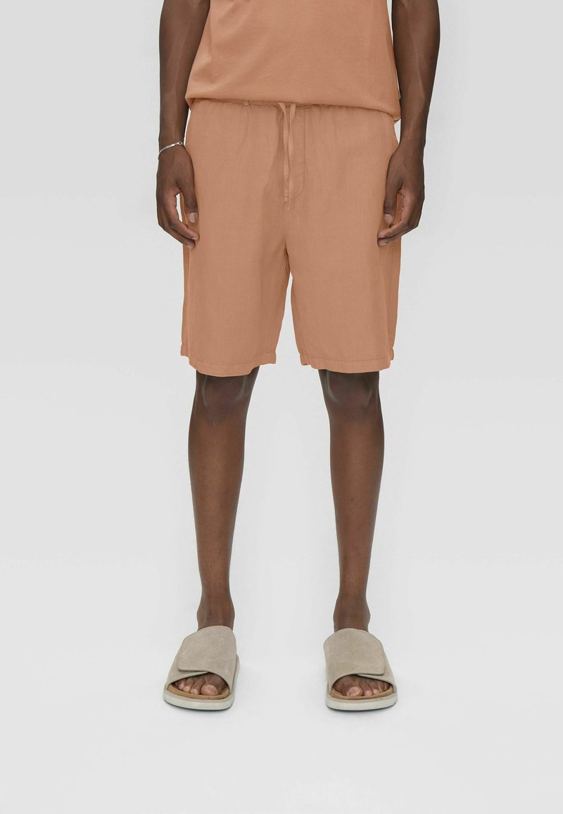 Homme portant un short marron clair avec cordon de serrage et des sandales taupe sans bride, debout devant un fond clair uni.