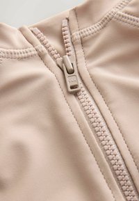 Beežist kangast, millel on sile tekstuur. Lukul on plastikust tõmbelukk ja nähtavad õmblused. Materjal tundub veniv ja kerge.