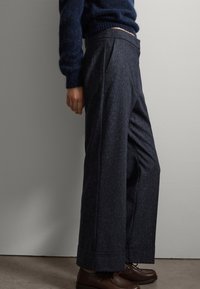 Brede broekspijpen in een donkere, getextureerde stof die een subtiel gestructureerd patroon vertoont, met een hoge taille en nette plooien. Gepaard met bruine schoenen.
