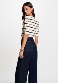 Gestreifter, cremefarbener Strickoberteil mit kurzen Ärmeln, kombiniert mit hoch taillierten, dunkelblauen Jeans im weiten Bein. Auffällige goldene Hardware-Details bei den Accessoires.