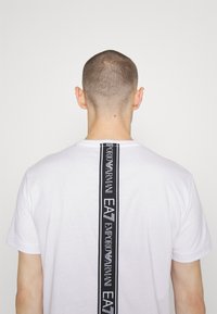Camiseta de algodón blanca con una franja de logo vertical negra en la parte posterior, con un patrón de texto "Emporio Armani EA" en diseño repetido.