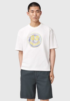 Mies, jolla on valkoinen lyhythihainen t-paita, jossa keltainen ja sininen hymyilevä kasvokuva, sekä tummanharmaat shortsit, seisoo yksinkertaisen taustan edessä.