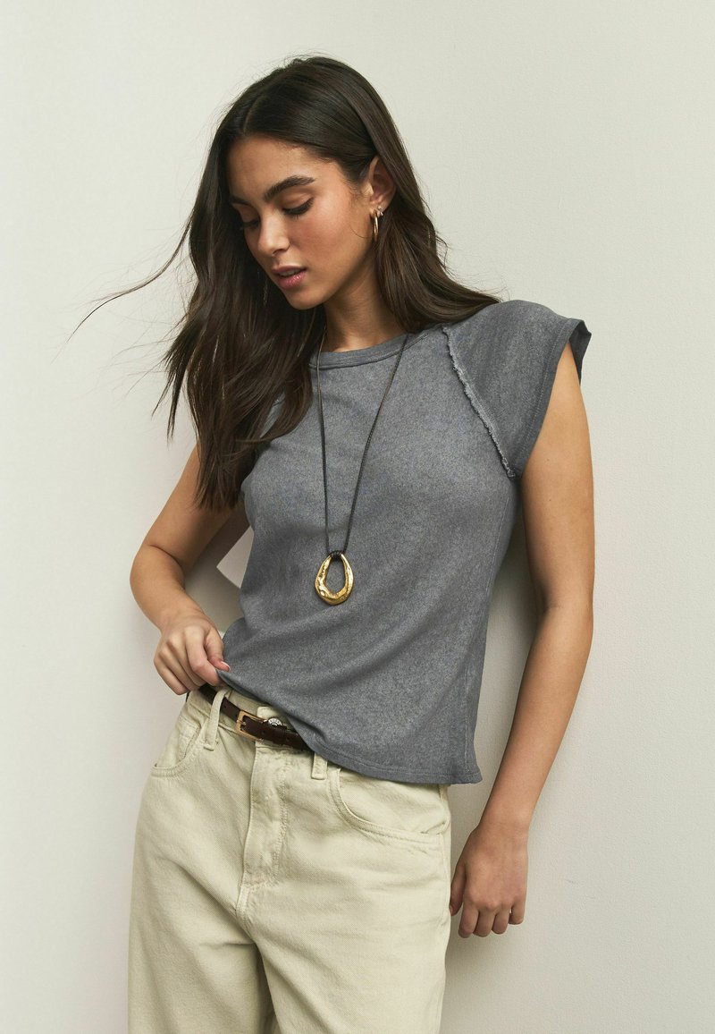 Haut gris à manches courtes avec un ourlet légèrement effiloché, associé à un pantalon beige et un collier pendentif en or. Tissu décontracté et coupe ample.