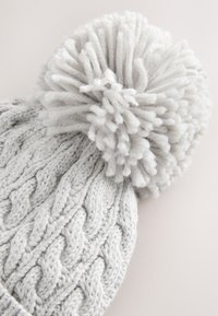 Strikket grå hue med et tykt, tekstureret kabelmønster og en stor, blød kvast på toppen. Blødt, varmt materiale, der er velegnet til koldt vejr.