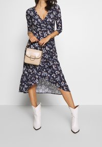 Robe portefeuille florale navy à manches trois-quarts, ourlet volanté, en tissu léger. Associée à un sac à main beige texturé et des bottes de cowboy blanches.