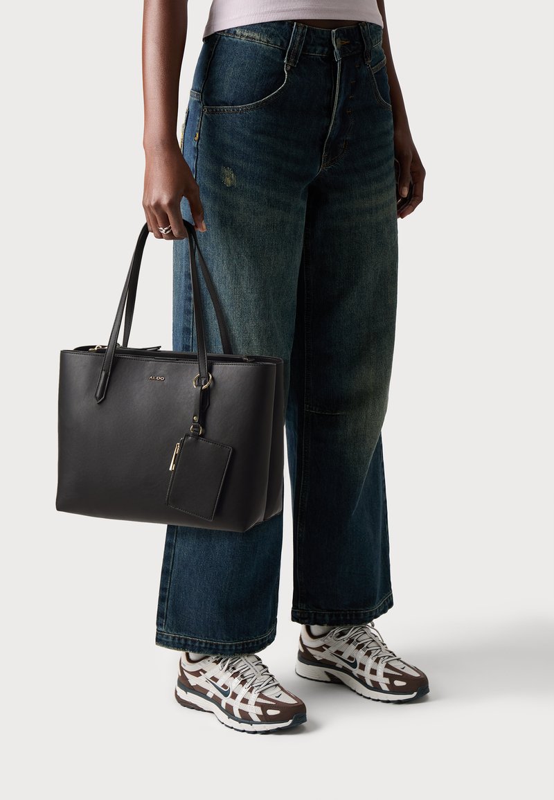 Schwarze Leder-Tasche mit zwei Griffen, goldenen Akzenten und einem abnehmbaren Etikett, kombiniert mit weit geschnittenen blauen Jeans und gemusterten Turnschuhen.