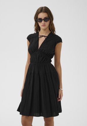 Femme portant une robe noire longueur genoux avec manches courtes, col en forme de trou de serrure avec lien, lunettes de soleil noires et bracelet argenté, debout sur fond blanc.