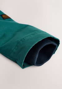 Rukáv tyrkysovej bundy s tmavomodrým rebrovaným pleteným vnútorným lemom, ktorý vyčnieva z konca rukáva pre dodatočné teplo a pohodlné priliehanie.