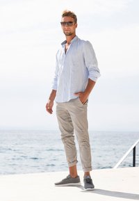 Helles blaues Leinen-Hemd mit Knöpfen, beige hochgekrempelte Chinos, graue Sneaker, steht am Wasser unter klarem Himmel. Sonnenlicht beleuchtet die Szene.