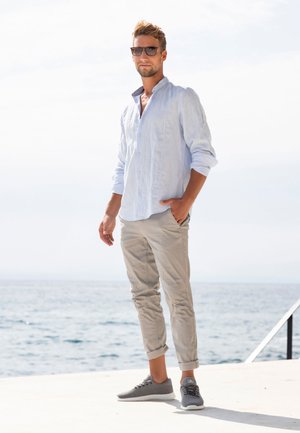 Light blue linen button-up shirt, beige rolled-up chinos, gray sneakers, standing by water with a clear sky. Sunlight illuminating the scene.