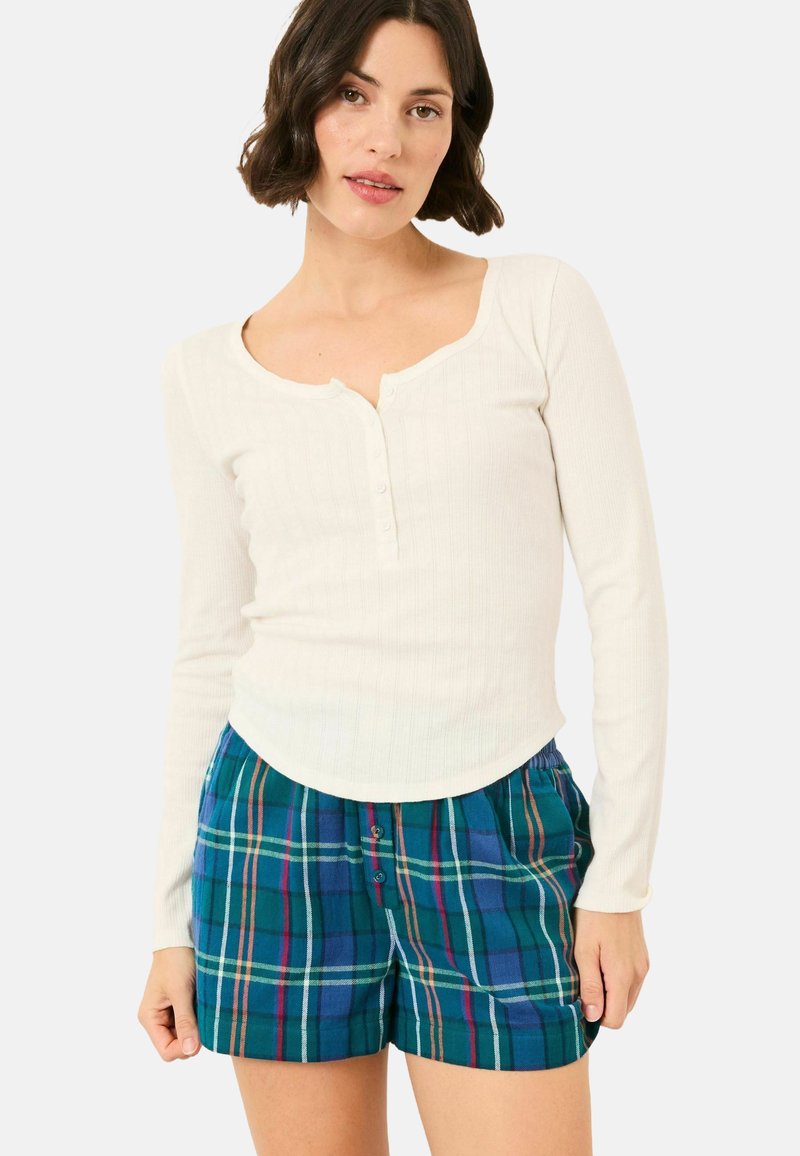Woman wearing a white long-sleeve ribbed top with buttons and blue-green plaid shorts standing against a plain background.