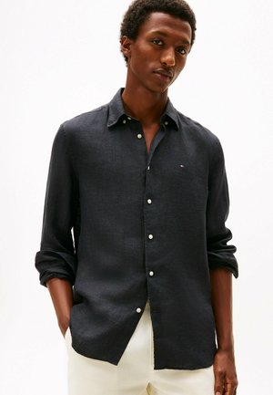 Jeune homme portant une chemise foncée boutonnée avec les manches retroussées et un pantalon clair, debout devant un fond blanc uni.