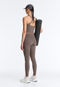 Žena v sivom športovom oblečení a šiltovke drží zrolovanú jogovú podložku, stojaca chrbtom k divákovi na jednoliatom bielom pozadí.