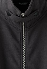 Veste zippée gris foncé avec un col haut, des boutons-pression et une fermeture éclair métallique visible, marque Stradivarius, fabriquée au Bangladesh.