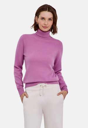 Femme portant un pull à col roulé violet et un pantalon blanc à cordon, les mains dans les poches, regardant devant avec un léger sourire.