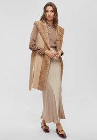 Gilet in pelliccia sintetica beige sopra una blouse marrone e una gonna in satin beige, con una texture liscia e un design al ginocchio. Scarpe piatte marroni completano l'outfit.