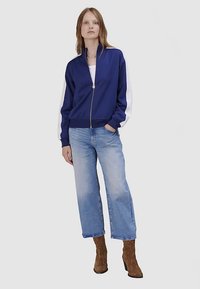 Veste zippée bleu marine avec manches blanches, en matériau doux. Associée à un jean large bleu clair et des bottes à talons marron.