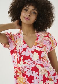 Cvetna obleka z živahnimi rdečimi, roza in rumenimi odtenki. Ima V-izreze in kratke rokave; tkanina je gladka in ima cvetlični vzorec.