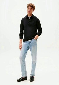 Schwarzes langärmliges Polohemd mit kleinem Logo, hellblaue gerade geschnittene Jeans und schwarze Slipper, stehend vor einem neutralen Hintergrund.