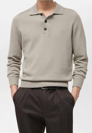 Mann trägt einen hellbeigen gestrickten Langarm-Polopullover, in dunkelbraune Hose gesteckt, mit Händen in den Taschen.