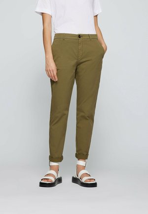 Mannequin portant un pantalon olive retroussé, une chemise blanche à manches courtes et des sandales à plateforme blanches avec semelles noires, debout devant un fond uni.