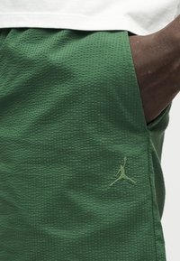 Main d'une personne à la peau foncée dans une poche de short vert texturé avec un logo Jumpman brodé, portant une chemise blanche au-dessus.