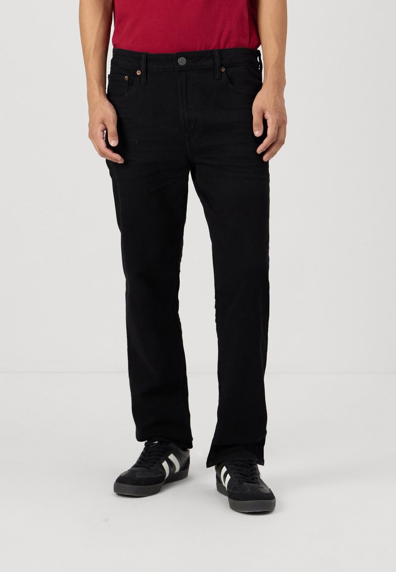 Jeans de denim negro con corte recto, cinco bolsillos y detalles de costura sutiles, combinados con una camiseta roja y zapatillas negras.