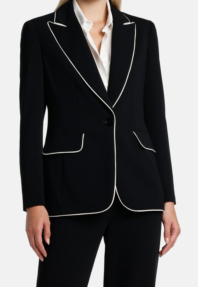 Femme portant une veste de costume noire avec un liseré blanc le long des revers, des poches et des bords, par-dessus une chemise blanche boutonnée.